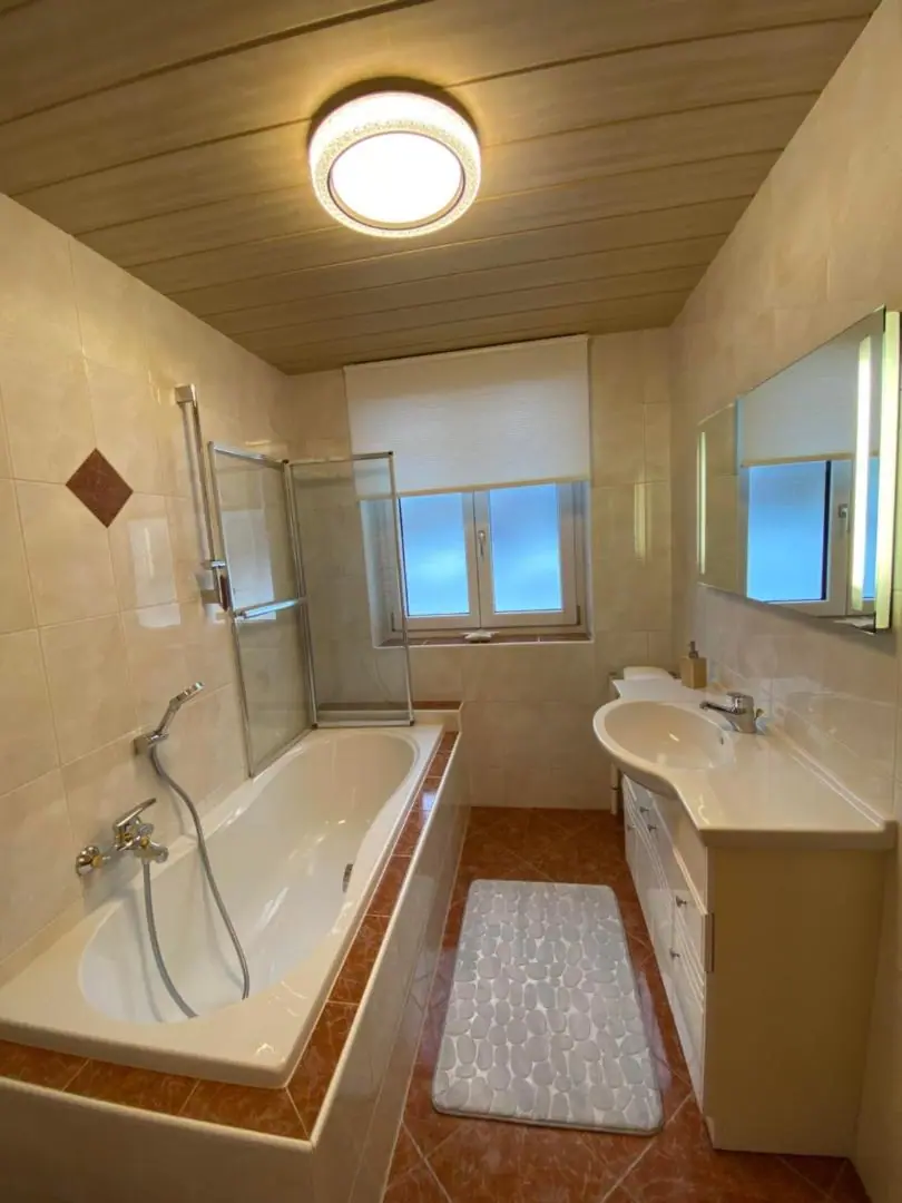 Badezimmer mit Badewanne samt Duschwand, Waschtisch, beleuchtetem Spiegel und Fenster