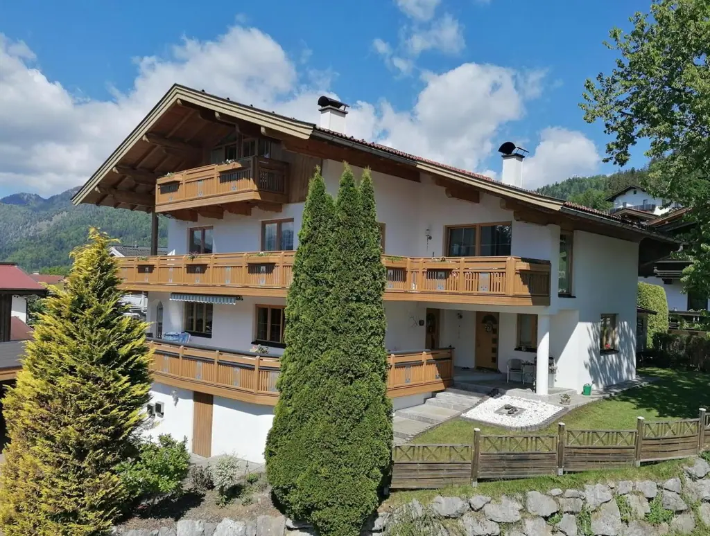 Das Haus der Familie Achhorner im Sommer: weiß verputzt, mit Holzbalkonen, Garten und zwei hohen Thujen, dahinter bewaldete Berge