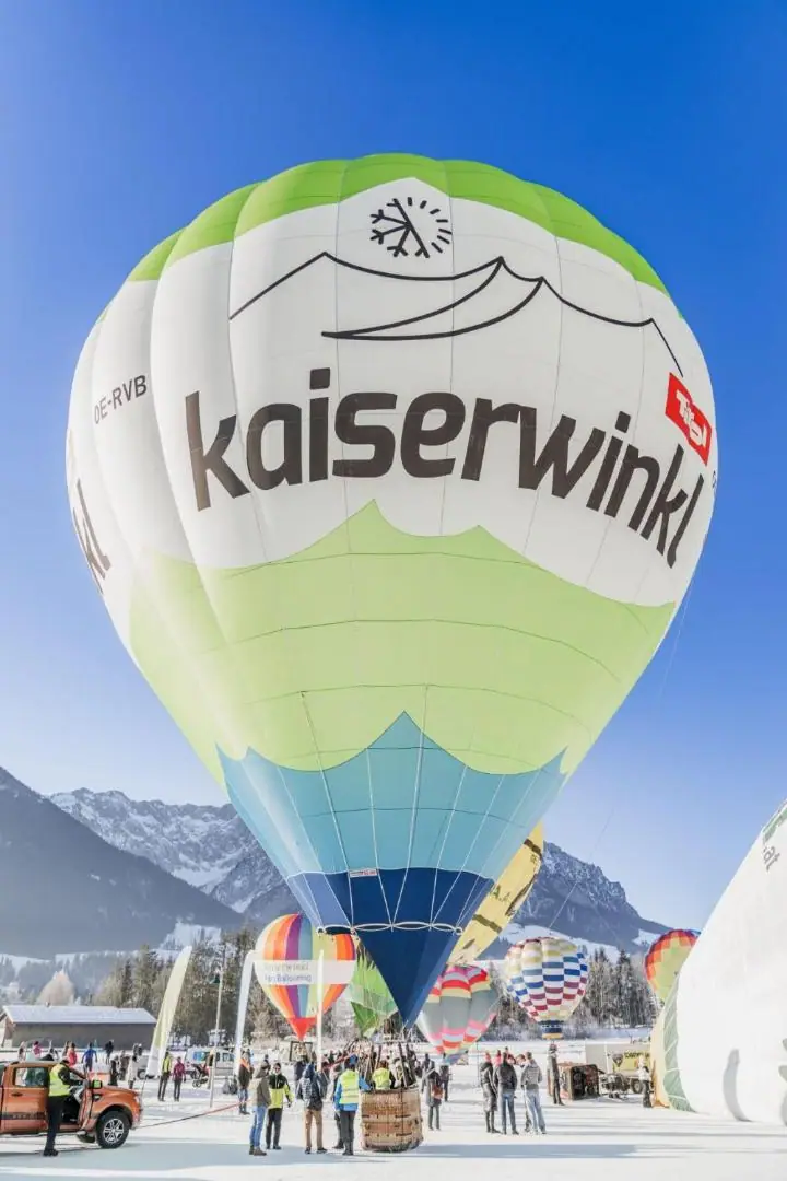 Heißluftballon mit Kaiserwinkl-Schriftzug beim Start im Schnee, dahinter weitere Ballone und Berge