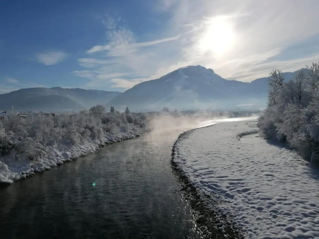 Fluss im Winter mit verschneiten Ufern, Morgendunst und Bergen im Hintergrund