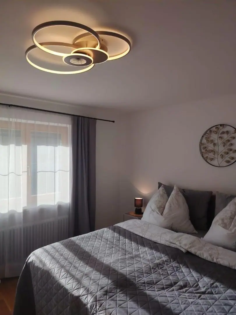 Schlafzimmer mit Doppelbett, grauer Steppdecke, Fenster mit Vorhängen und goldener Ring-Deckenleuchte