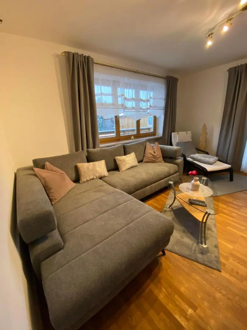 Wohnzimmer mit großem grauem Ecksofa, Glastisch, Parkettboden und breitem Fenster
